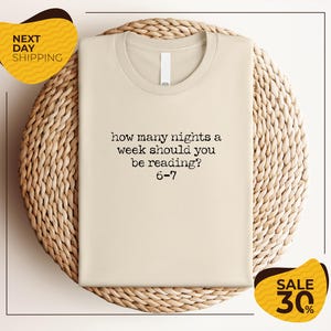 A beige t-shirt with a humorous text printed on it, placed on a woven basket or mat.
