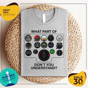 a gray t-shirt with a graphic design depicting various flight instruments and the text "WHAT PART OF DON'T YOU UNDERSTAND?" The t-shirt is displayed on a woven placemat, and there are two yellow circular stickers in the image.