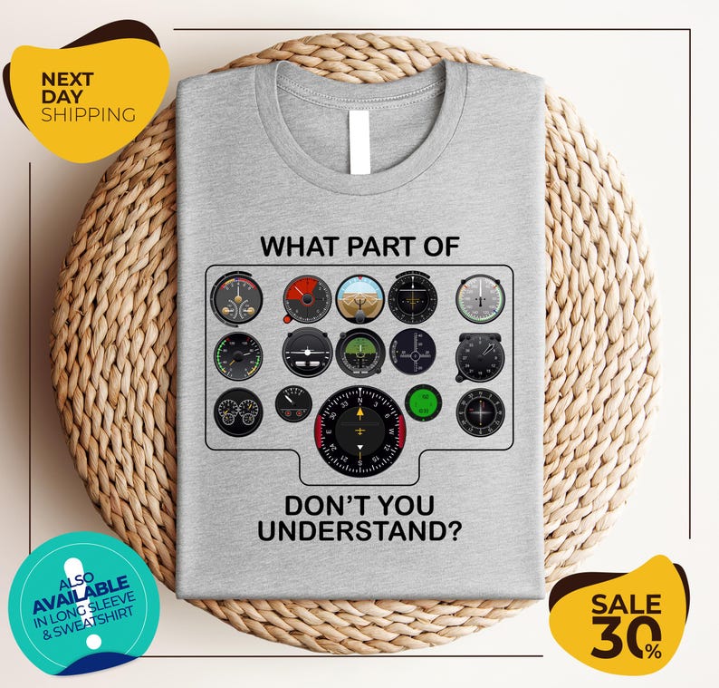a gray t-shirt with a graphic design depicting various flight instruments and the text "WHAT PART OF DON'T YOU UNDERSTAND?" The t-shirt is displayed on a woven placemat, and there are two yellow circular stickers in the image.