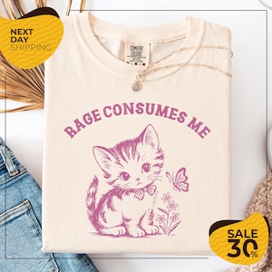 a white t-shirt with a graphic design of a cat and a butterfly, along with the text "RAGE CONSUMES ME" printed on it. The t-shirt is displayed on a white background, and there are some jeans and a sweater visible in the background.