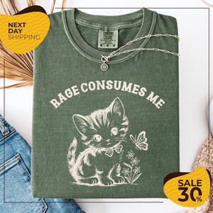 a green t-shirt with a white graphic design of a cat and flowers, along with the text "RAGE CONSUMES ME" printed on it. The t-shirt is displayed on a white background, and there are yellow and black sale signs indicating a 30% discount.
