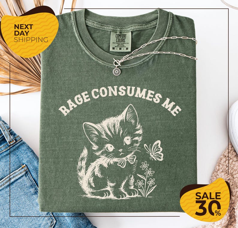 a green t-shirt with a white graphic design of a cat and flowers, along with the text "RAGE CONSUMES ME" printed on it. The t-shirt is displayed on a white background, and there are yellow and black sale signs indicating a 30% discount.