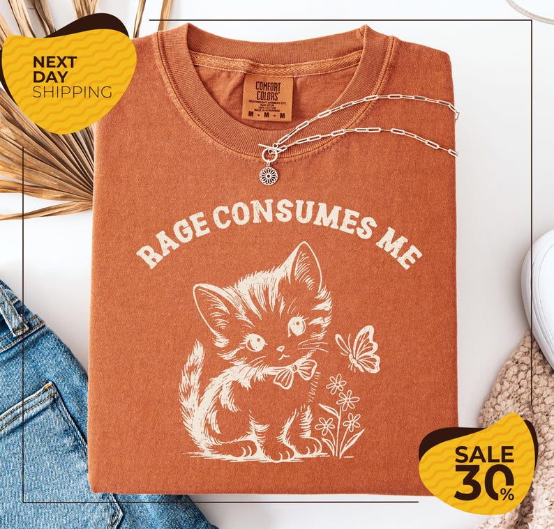 a red t-shirt with a white cat and flowers printed on it, along with a necklace and a butterfly. The t-shirt is displayed on a white background, and there are yellow and black sale signs in the background.