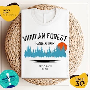 a white t-shirt with a graphic design of a forest scene, along with text indicating the location as Viridian Forest National Park. The t-shirt is displayed on a woven basket, and there are two yellow circular stickers in the image.