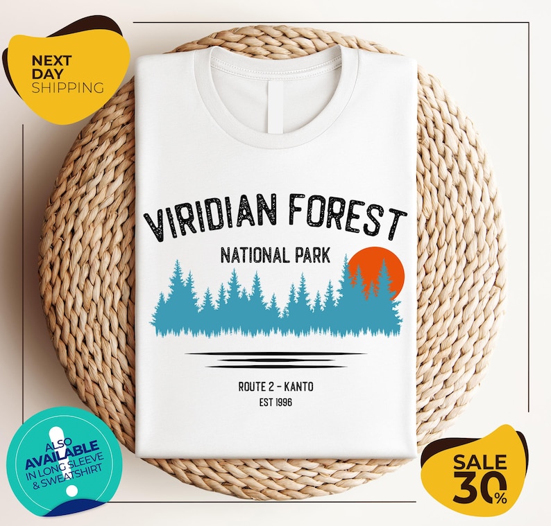 a white t-shirt with a graphic design of a forest scene, along with text indicating the location as Viridian Forest National Park. The t-shirt is displayed on a woven basket, and there are two yellow circular stickers in the image.