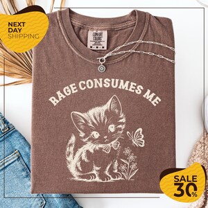 a brown t-shirt with a white cat and flowers printed on it, along with a necklace and a chain. The t-shirt is displayed on a white background, and there are yellow and black sale signs in the image.
