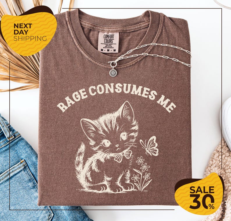 a brown t-shirt with a white cat and flowers printed on it, along with a necklace and a chain. The t-shirt is displayed on a white background, and there are yellow and black sale signs in the image.