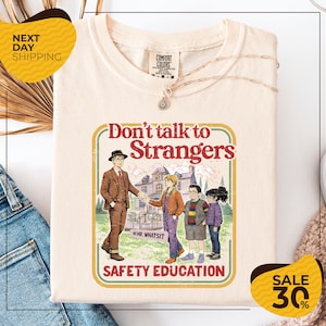 May include: Cream-colored t-shirt with a graphic that reads "Don't talk to Strangers" in a retro style. The graphic features an illustration of a man and children. The shirt also has the text "SAFETY EDUCATION".