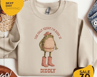 May include: Beige sweatshirt with a cartoon frog wearing a cowboy hat and boots. The text "THE ONLY SQUAT I'M DOIN IS DIDDLY" is printed above and below the frog illustration. The sweatshirt has a crew neck and long sleeves.