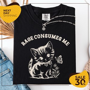 a black t-shirt with a white graphic design depicting a cat and flowers, along with the text "RAGE CONSUMES ME" printed on it. The t-shirt is displayed on a white background, and there are jeans and a necklace visible in the background.