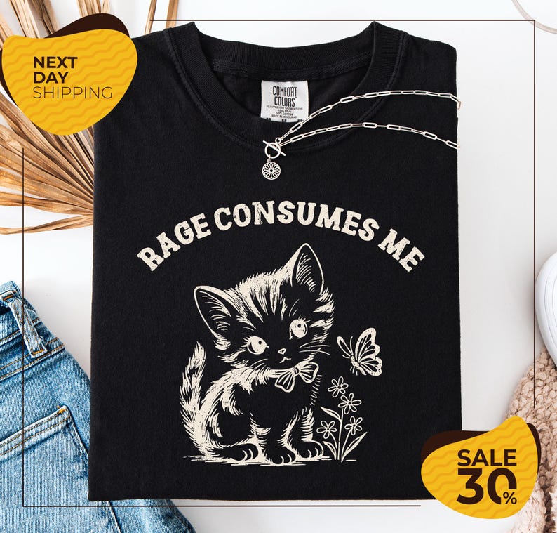 a black t-shirt with a white graphic design depicting a cat and flowers, along with the text "RAGE CONSUMES ME" printed on it. The t-shirt is displayed on a white background, and there are jeans and a necklace visible in the background.