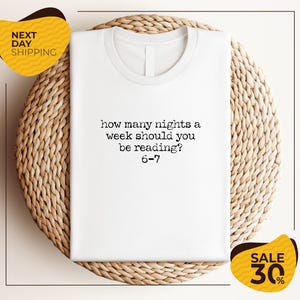 A white t-shirt with a humorous text printed on it, placed on a woven placemat. The text reads "how many nights a week should you be reading 6-7". The image also includes two yellow sale signs, one for "NEXT DAY SHIPPING" and the other for "SALE 30%".