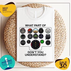 a white t-shirt with a graphic design depicting various flight instruments and the text "WHAT PART OF DON'T YOU UNDERSTAND?" The t-shirt is displayed on a woven placemat, and there are two yellow circular stickers with text on them.