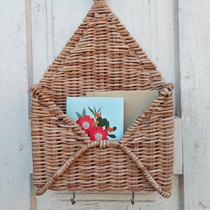 May include: A woven wicker wall organiser shaped like an envelope with three hooks on the bottom. The organiser is hanging on a white wooden wall and has two cards inside.