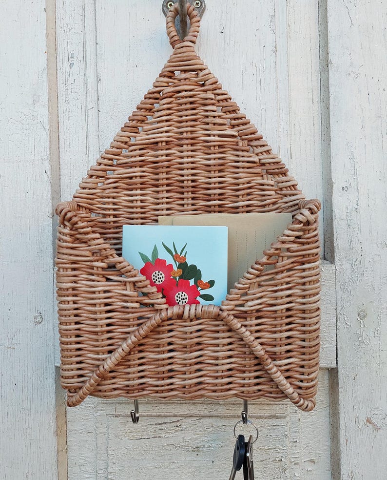 May include: A woven wicker wall organiser shaped like an envelope with three hooks on the bottom. The organiser is hanging on a white wooden wall and has two cards inside.