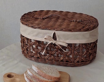 Op de afbeelding: Een bruine rieten broodmand met een beige stoffen deksel en een wit lint. De mand is ovaalvormig en heeft een geweven patroon. Er liggen vier sneetjes brood op een houten snijplank voor de mand.