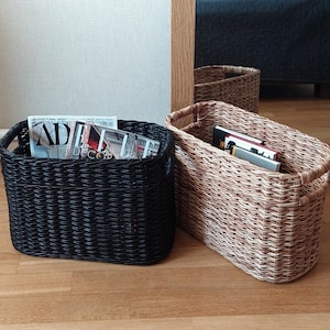 May include: Two woven storage baskets, one black and one natural brown, with handles. The black basket is filled with magazines, including one with the text "AD" and "DECOR".