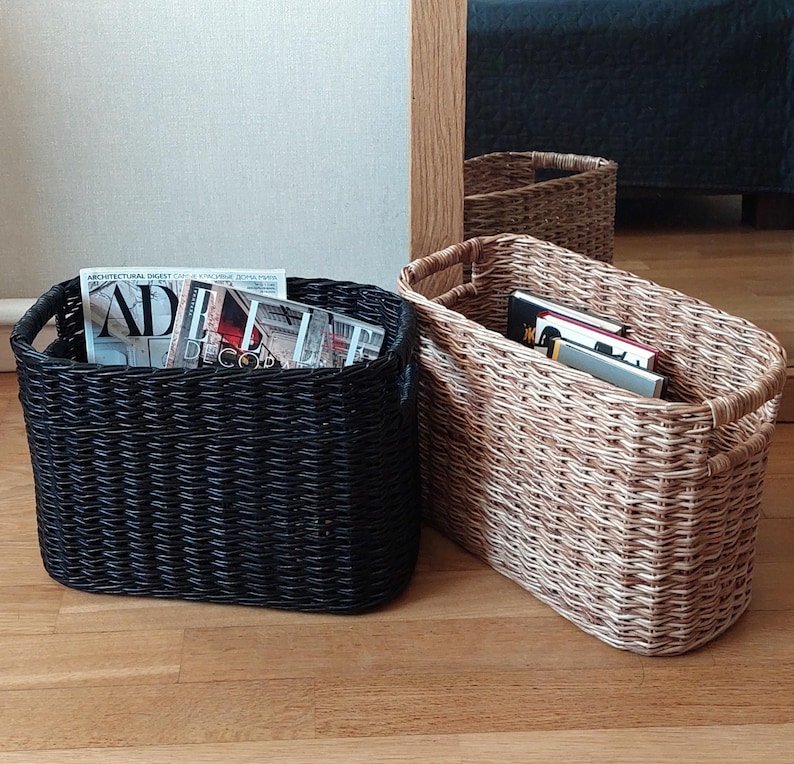 May include: Two woven storage baskets, one black and one natural brown, with handles. The black basket is filled with magazines, including one with the text "AD" and "DECOR".