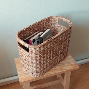May include: A woven wicker basket with two handles, filled with books. The basket is light brown and has a natural woven pattern. The books are stacked vertically and have different coloured covers.