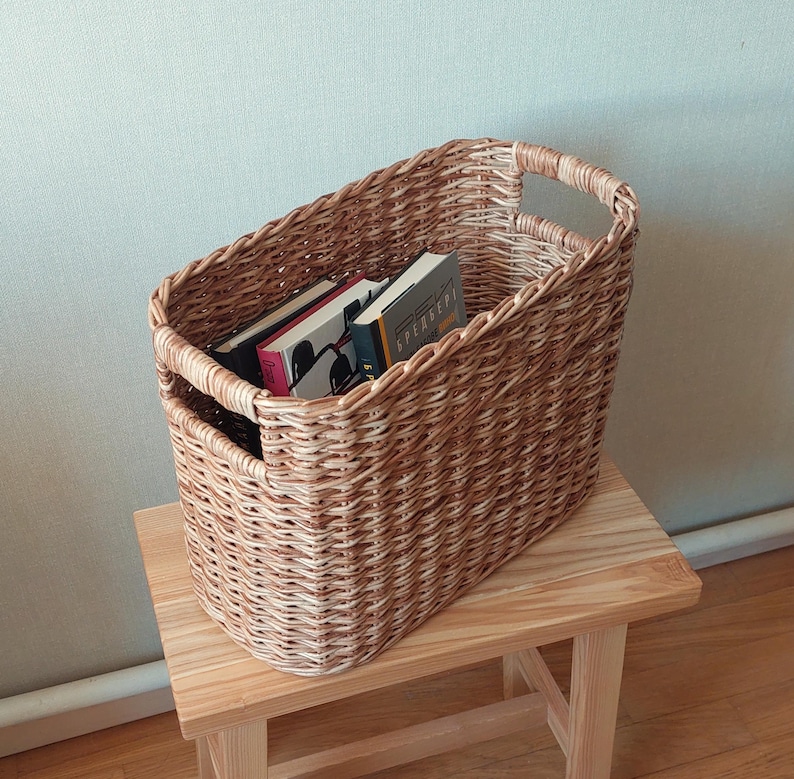May include: A woven wicker basket with two handles, filled with books. The basket is light brown and has a natural woven pattern. The books are stacked vertically and have different coloured covers.