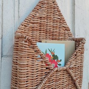 May include: A woven wicker wall organiser shaped like an envelope with two hooks on the bottom. The organiser has a light brown colour and is holding two cards with floral designs.
