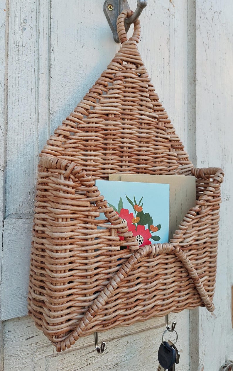 May include: A woven wicker wall organiser shaped like an envelope with two hooks on the bottom. The organiser has a light brown colour and is holding two cards with floral designs.