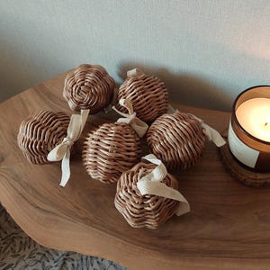 May include: Six woven rattan spheres with cream-colored ribbon loops are arranged on a wooden surface. A lit candle in a glass jar sits to the right. The spheres are a light brown color and have a textured surface.