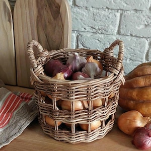 Könnte beinhalten: Ein geflochtener Korb aus Weide mit zwei Griffen, gefüllt mit roten Zwiebeln und Knoblauch. Der Korb steht auf einer Holzoberfläche mit anderen Zwiebeln und einem Kürbis.