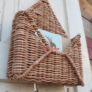 May include: A woven wicker wall-mounted mail holder with two hooks underneath. The holder is shaped like an envelope and has a light brown colour.