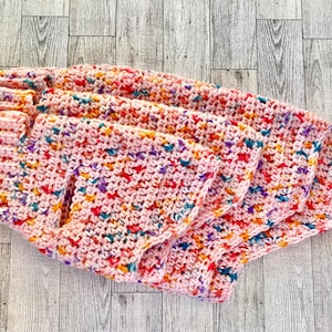 May include: A pink crocheted blanket with colorful flecks of red, yellow, blue, and purple. The blanket is folded and appears soft and textured, likely handmade. The yarn has a speckled appearance.