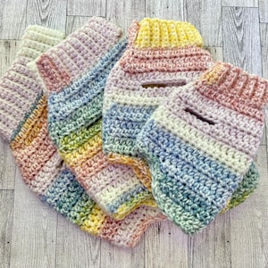 May include: A collection of four handmade crochet sweaters for pets, each featuring a variegated pastel yarn pattern in shades of soft pink, yellow, blue, and cream. The garments have a ribbed collar and a small rectangular opening on the back.