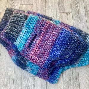 May include: A thick, hand-crocheted garment featuring a variegated yarn pattern in shades of deep blue, purple, magenta, and bright turquoise. The piece has a ribbed texture, a central vertical opening, and a chunky, multi-colored knit construction.