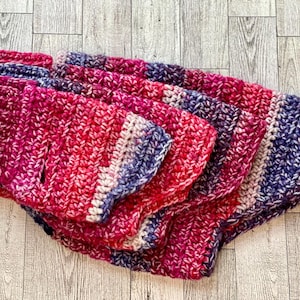 May include: A stack of four handmade crochet dog sweaters in varying sizes. The sweaters are primarily red and pink with blue and white accents. The yarn has a textured appearance.
