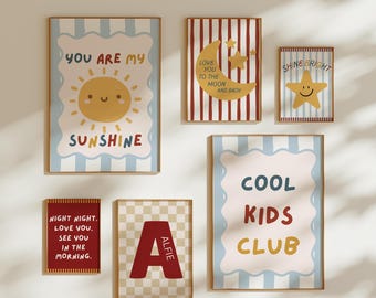 May include: A collection of framed art prints with playful designs. The prints feature phrases like "You are my sunshine" and "Cool Kids Club," along with a smiling sun, moon, and stars. The color palette includes blue, red, and beige.