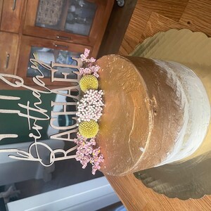 Könnte beinhalten: Eine goldene und weiße Torte mit einem Holzaufsatz, auf dem "Happy Birthday Rachael" steht. Die Torte ist mit rosa und gelben Blumen verziert.