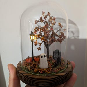 Op de afbeelding: Een decoratieve glazen koepel met een Halloween-thema. De miniatuurscène bevat een boom met herfstbladeren, een spook, pompoenen, een zwarte kat en een grafsteen op een bed van bladeren. Een kleine lantaarnpaal verlicht de scène.