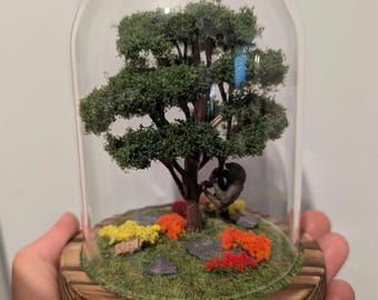 Könnte beinhalten: Ein Miniatur-Baumdiorama in einer Glaskuppel. Die Szene zeigt einen detaillierten Baum mit grünen Blättern, einen kleinen Affen, bunte Blumen und einen Holzsockel. Die Kuppel wird in einer Hand gehalten.