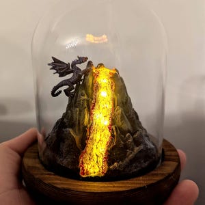Könnte beinhalten: Eine Miniatur-Drachenfigur, die auf einem kleinen, felsigen Berg mit einem glühenden orangefarbenen Lavastrom in einer Glaskuppel auf einem Holzsockel sitzt.