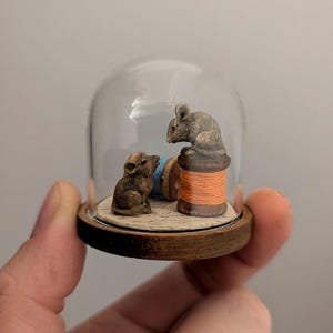 Puede incluir: Dos pequeñas figuritas de ratones marrones están dentro de una cúpula de vidrio con una base de madera. Un ratón está sentado en un carrete de hilo naranja, y el otro ratón está sentado en un carrete de hilo azul.