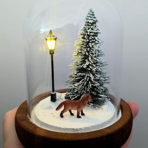 May include: A miniature winter scene inside a glass dome. The scene includes a small brown fox, a snow-covered evergreen tree, and a lit street lamp. The dome sits on a wooden base.