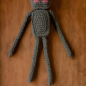 May include: A handmade grey crochet doll with a rectangular body, long arms and legs, and a square head. The doll has pink accents for the eyes. The doll is displayed on a wooden surface.