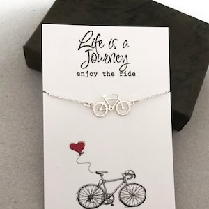 May include: Silver necklace featuring a bicycle pendant and the text "Life is a Journey, enjoy the ride". The necklace is presented on a white card with a red heart balloon and a bicycle illustration.