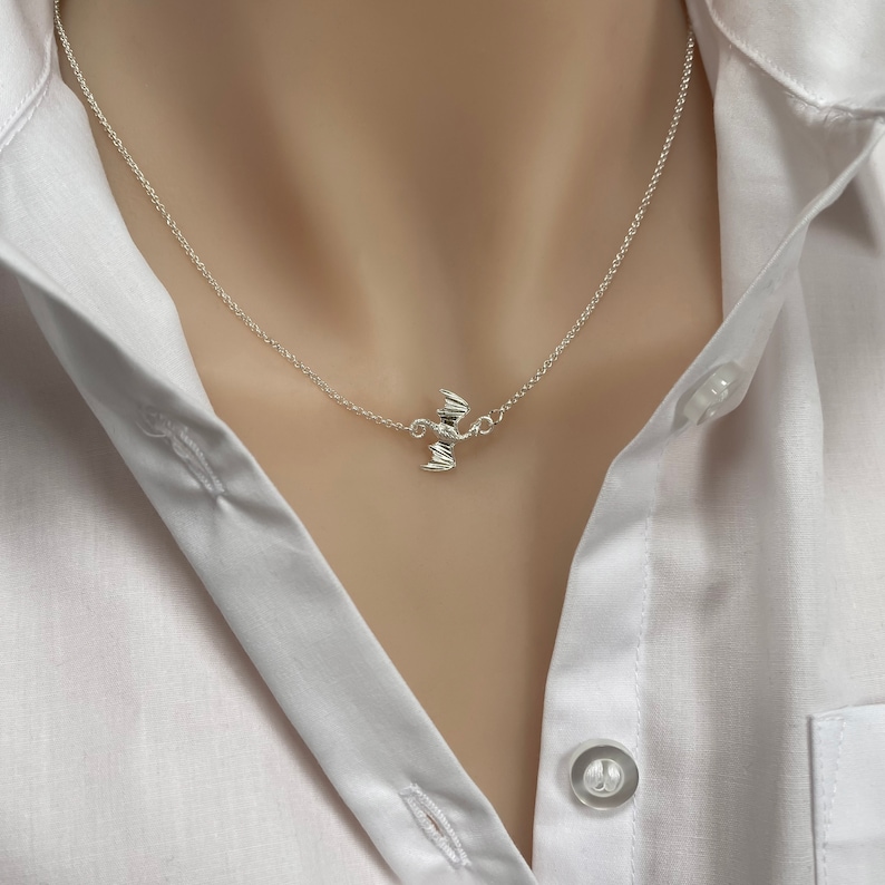 May include: A silver necklace with a small, detailed bat pendant. The necklace is on a model wearing a white shirt.