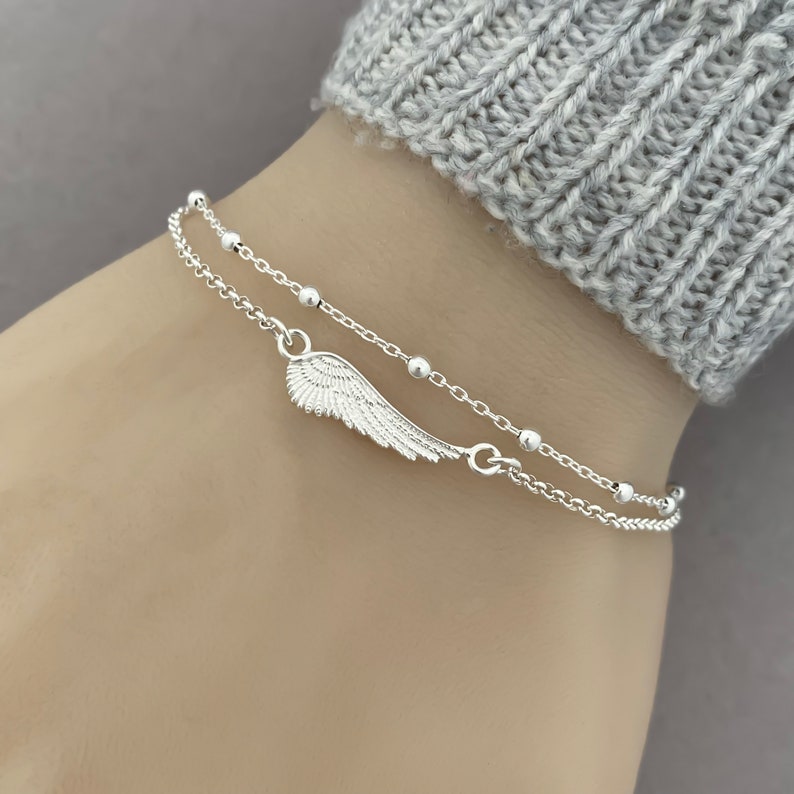 Puede incluir: Un brazalete de cadena de plata con un delicado dije de ala de &aacute;ngel y peque&ntilde;as cuentas de plata. El brazalete est&aacute; formado por dos cadenas superpuestas.
