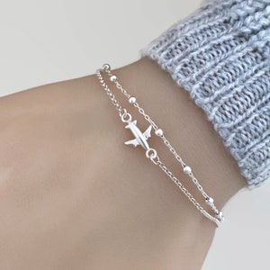 May include: A silver bracelet featuring a small airplane charm and a delicate chain with small beads. The bracelet is layered on a person's wrist.