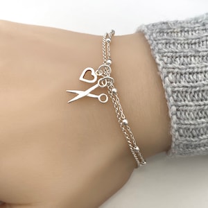 May include: A silver chain bracelet with a heart and scissors charm. The bracelet is on a person's wrist.