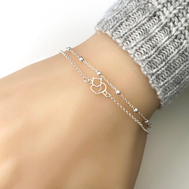 May include: A silver chain bracelet with a small cat charm. The bracelet is layered with a second chain with small silver beads.