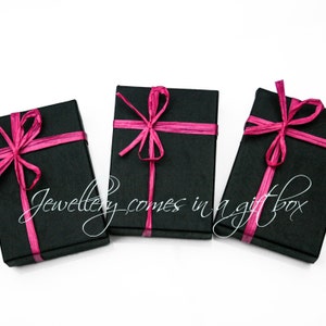 May include: Three black gift boxes with pink ribbon bows. The boxes are on a white background.  "Jewellery comes in a gift box" is written in white cursive on the image.