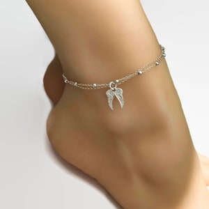 May include: Silver ankle bracelet with a delicate chain and small beads. A charm in the shape of angel wings hangs from the chain.