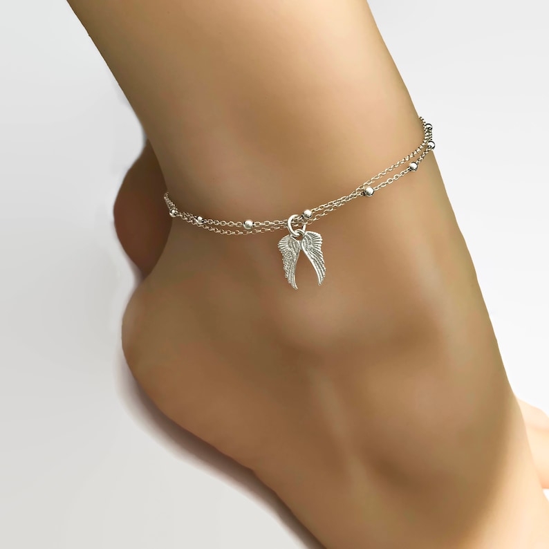 May include: Silver ankle bracelet with a delicate chain and small beads. A charm in the shape of angel wings hangs from the chain.